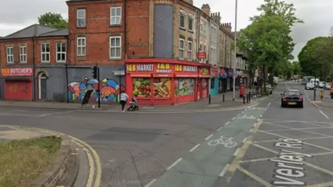 Google A junction on a main shopping street. A mini market, with red frontage, is on a corner. Next door, is a shop shutter covered in colourful graffiti. 