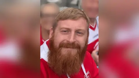 Humberside Police Family photograph of James Adam Barlow. He has red hair and a beard and is wearing a red and white Hull Kingston Rovers top.
