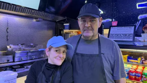 The photo shows two people, one of them being Darren Clark, smiling at the camera. They're both wearing dark-coloured tops, and black aprons. They're both wearing baseball caps. They're standing in a café. To the right of the photo is a fridge filled with cans of pop, and chocolate bars. To the right there are metal grills and fryers.