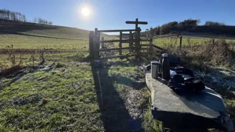 BBC A view of a footpath with the sun low in the sky peeping up behind a hill. There are green fields up ahead and in the foreground is a bench with flasks, gloves and walking stick. 