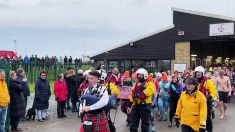 BBC Swimmers in various outfits march out of the lifeboat station behind a bagpiper in traditional Scottish dress