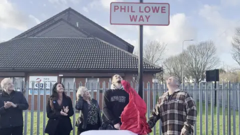 BBC Five people stand underneath a road sign. It is in red letters against a white background and says Phil Lowe Way. Underneath are members of his family, including his son, who has just pulled down a red sheet that was covering the sign.