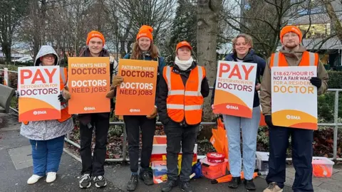 Sarah Sanderson/BBC Five people standing outside Hull Royal Infirmary wearing orange beanie hats and holding placards. One reads: "Pay restoration for doctors". Another says: "Patients need doctors, doctors need jobs".