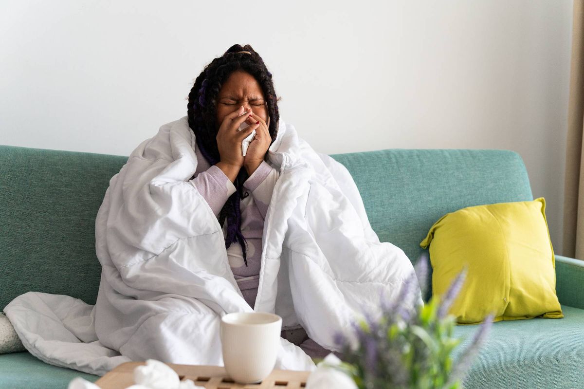 Young woman sitting on the sofa at home, covered with a blanket, blowing her nose, suffering from a cold or flu