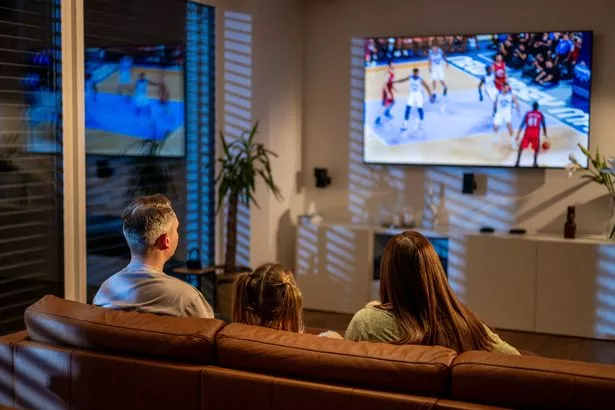 A Caucasian family, including a man and two women, relaxes on a sofa while watching an intense basketball game on TV in a modern, warmly-lit living room.