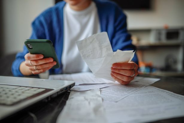 middle-aged woman with glasses, laptop, smartphone, and papers at a table at home