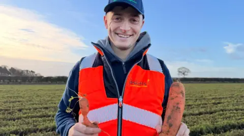 BBC A man with a blue coat and blue cap is standing in a field holding a large carrot in one hand and a deformed carrot in another. He also has an orange hi-vis vest on. He is smiling and looking into the camera.