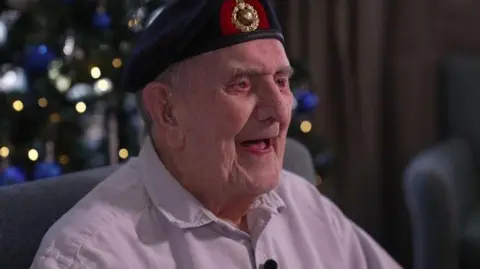 BBC An elderly veteran wearing a navy beret with a green and gold logo at the front. He is smiling with his mouth open and wearing a long-sleeved light pink shirt. He is sat in a grey chair and the background is blurred but you can see fairy lights from a Christmas tree.