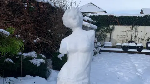 Emily Bates A classical Greek style sculpture of a woman with no arms made from snow. Behind the sculpture is a snowy suburban garden.