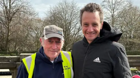 Jonathan Reed/BBC An older man wearing a navy fleece, a fluorescent yellow jacket and a running cap standing beside a taller, younger man, with short curly brown-grey hair and wearing a black Adidas coat.