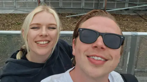 Lewis Budden A woman with blonde plaits wearing a black hoodie and a man with slicked back light brown hair wearing a white T shirt post for a selfie in front of a festival stage.