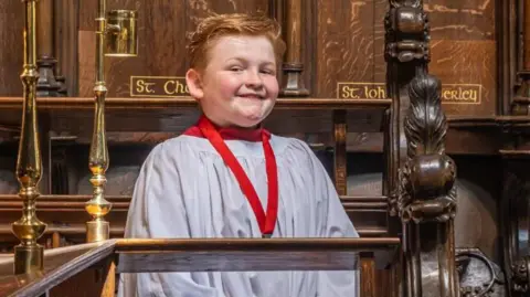 Helen Tabor Photography Henry Burr dressed in a chorister gown wearing a red medal smiling in a church 