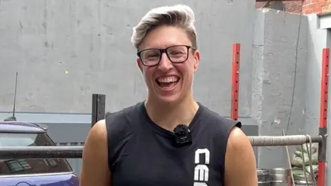 Laura Hollywood stands outdoors in front of a grey wall, wearing a sleeveless black shirt with white vertical text on the front.