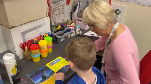 BBC / Grace McGrory The photo shows a young boy who is reading to an older woman. He has short brown hair and a blue sweatshirt. She has short, blonde hair and is wearing a pale pink jumper. They're sat at a table in a classroom. There are scissors, playdough pots, and various folders and bits of paper on the desk. 