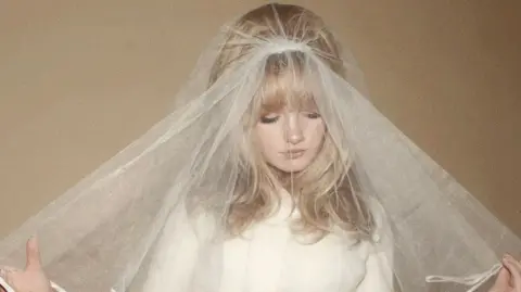 Aimée Fatale A blonde haired woman wearing a white dress and a bridal veil looks towards the ground.