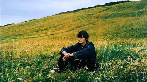 Zander A brown haired man wearing a denim jacket and jeans sits in a grassy field.