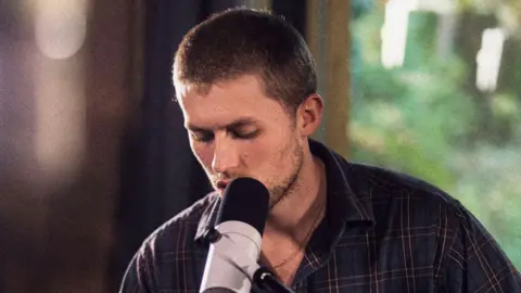 Henri Gaston A man with short brown hair singing into a microphone. He is wearing a checked shirt.