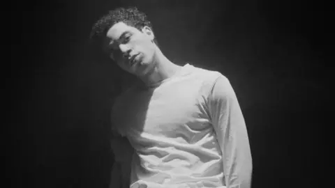 Kenzo Jae A black and white image of a man in a long sleeved white top staring at the camera. He has short, curly, dark hair.