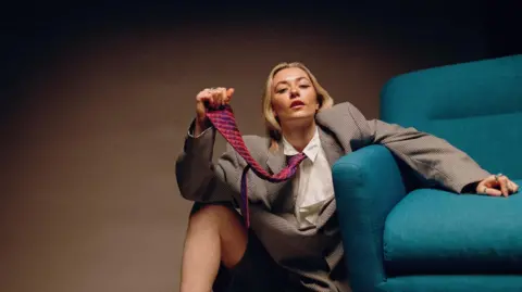 Lois A blonde haired woman in a sports jacket, red and blue tie and white shirt sits on the floor next to a blue chair.