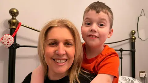 BBC A woman smiles as she sits on a bed with her son, who has his arms around her. The woman has strawberry-blond hair, a silver nose stud and wears a black T-shirt. The boy has short blond hair and wears a red T-shirt. The bed has a black and gold frame. The wall is cream-coloured.