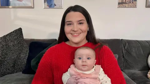 Keira sits on the sofa wearing a bright red jumper and holding her baby daughter in her arms as they both look at the camera.