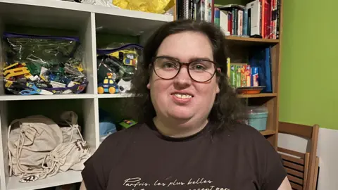Kirsty has brown shoulder length wavy hair and is wearing glasses and a brown T-shirt. She is standing in front of bookcases which are displaying toys and books. 