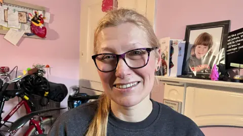 BBC Monika sits in front of her fireplace and a photograph of her son and smiles at the camera wearing a grey jumper and dark purple glasses.
