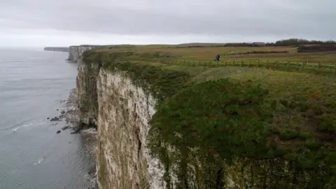 A sweeping view of Bempton Cliffs on the East Yorkshire coast. Towering chalk cliffs dominate the foreground, their sheer white faces streaked with grey and green vegetation. At the top, a grassy plateau stretches inland, enclosed by a wooden fence that runs along a narrow path. A single person is visible walking along the path, adding scale to the vast landscape. Beyond the cliffs, the coastline continues in a series of rugged headlands fading into the distance. Below, the North Sea appears calm and grey under an overcast sky, with soft waves breaking against the rocky base of the cliffs. The overall scene conveys a dramatic, windswept coastal environment. 