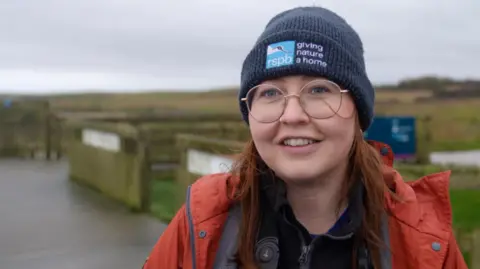 BBC A woman wearing a dark blue beanie hat and orange waterproof jacket stands on a wooden boardwalk at a nature reserve. Binoculars hang around her neck. Behind her are grassy fields, a wooden fence, and a winding path under an overcast sky.