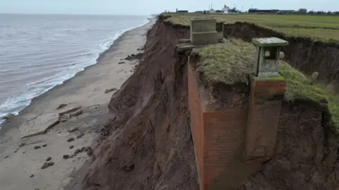 A large brick structure, mainly underground, is sticking out of the side of a clay cliff on a fast-eroding coastline. The bunker has two concrete structures, one a large entrance, the other a squat chimney, above ground. The cliff is dark brown and topped with green grass. Green fields stretch off in the distance towards a village on the horizon. Beneath the cliffs, clay and other material has fallen on to a beach, with the grey sea lapping against it.