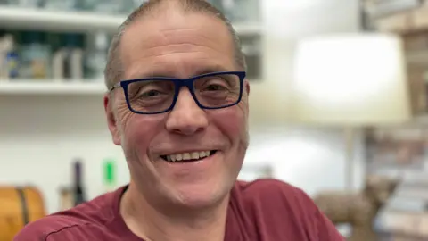 BBC A man smiles as he sits in a home in front of a lamp and a book shelf, which are out of focus. He wears glasses with dark-blue frames and a dark-red t-shirt.