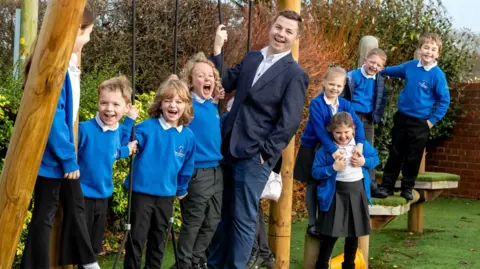 North Yorkshire Council Ben McAuley and a number of primary-age pupils stand on wooden children's play equipment, in a playground. Mr McAuley wears a navy blue suit and a white shirt. He has short brown hair. The pupils wear blue sweatshirts, black trousers or skirts and white polo shirts.