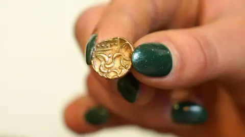 David Duggleby Auctioneers A small ancient gold coin with five dots across the top in a rectangular frame with an abstract depiction of a horse is being held up in a woman's hand who has nails painted in a glittery dark green.