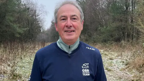 Dave Edwards/BBC Mark Burtonshaw faces the camera, dressed in athletic gear. He wears a light green thermal long-sleeved top, and navy blue t-shirt with a white GBR logo on the chest. He has short grey-white hair and stands against the backdrop of a frosty woodland.