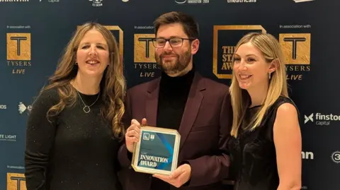 Hull Truck Theatre Hull Truck Theatre's executive director Janthi Mills-Ward, associate director Tom Saunders and head of marketing Laura Smith at the Stage Awards. Janthi Mills-Ward, who has long brunette hair, is smiling, wearing a black, long-sleeve glittery dress. Tom Saunders, who has short black hair and stubble, is holding an award. He is smiling, wearing glasses, a black turtle neck and a burgundy blazer. Laura Smith is wearing a black dress and has long, straight, blonde hair.
