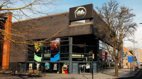 Tom Arran Photography The exterior of Hull Truck Theatre with black brick wall and large glass windows with the theatre's name on a sign at the top corner. Posters for upcoming plays hang in the window.