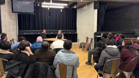 Hull Truck Theatre A group of GPs in a theatre sat on chairs facing away from the camera. They are looking at two people sat on chairs behind a table. 