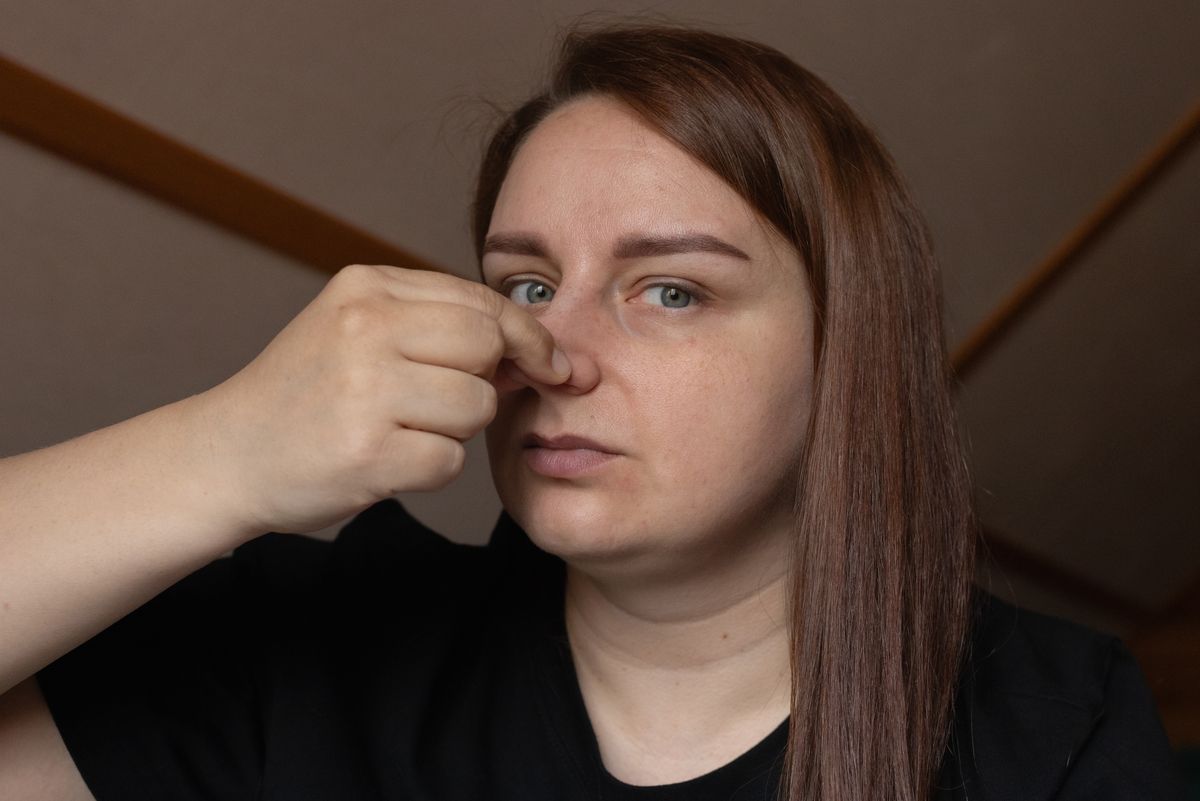 A woman indoors with long hair, pinching her nose due to an unpleasant odor. Her serious facial expression emphasizes an act of reaction or distaste. The setting appears domestic or casual.