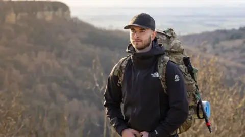 HOWARD KNIVETON A man stands in rural terrain. There are hills in the background. He wears a black jacket and baseball cap, and carries a large, military-style backpack.