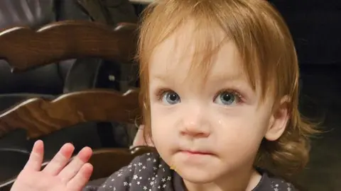 Story family A two-year-old girl with shoulder-length brown hair and blue eyes is wearing a dark top with white stars on it. She is sitting on a chair and is holding up her right hand.