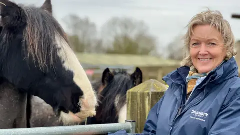 BBC/Linsey Smith Rachel Jenkinson wearing a navy coat stands to the right of the picture, a black and white horse to the left. Another smaller horse can be seen in the background.