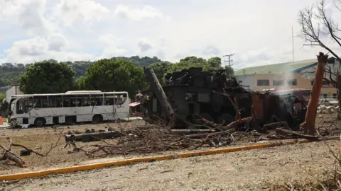 Miguel Gutierrez/EPA/Shutterstock Damage at an air base. The blackened remains of what appears to be a vehicle sits on scrubland beside a number of fallen and damaged trees. A burnt-out, white bus is parked to the left. A large building with sand-coloured walls and a green roof stands to the right. Trees and hills can be seen under a light blue, cloudy sky.