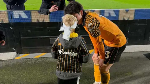 Submitted A footballer wearing a distinctive orange Hull City tiger striped kit speaks closely to a young child with bright blond hair. The little boy wears a white bandana and ear defenders.
