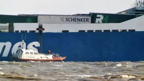 Michael Ludlow (MarineTraffic.com) A small boat is situated in what looks like choppy sea next to a port with a ladder suspended from it which goes up into a larger platform.