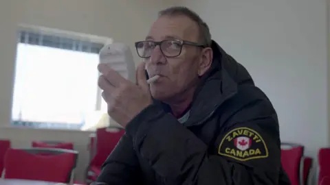 Eski / Hull City Council / SmokeFree Hull A screenshot from the 'Quitters' which shows Andy, a man who looks to be in his 60s, wearing glasses with a black frame. He is holding a small white machine in his hand, with a tube in his mouth as he blows in to it. He is white, has short grey hair and is wearing a black puffer coat with a Canada badge on the left sleeve. He is sitting in a room with red chairs and a large square window in the background. 