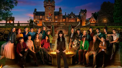 BBC/PA A large group photo of men and women all sat around a firepit. They are in front of a grand castle with floral bushes behind them. The group is split equally down the middle, half sat on either side of the firepit. In the middle stands a woman with long black hair and a full fringe- she looks directly at the camera whilst everyone else looks away. The sky behind them is a mixture of pinks and blues.