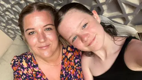 Lindsey Smith A woman and a girl pose for a picture with their heads tilted to the side. The woman has blonde hair plaited back off her face, she wears a pink, orange and yellow dotted dress and a thin gold necklace. The young girl has dark hair scraped back, and wears a one-strap black top. Behind them both is a detailed silver wall panelling.