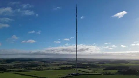 BBC The Belmont transmitter - a tall, thin transmission mast rises from the middle of bright green fields, supported by fine guy wires. Under a clear blue sky with scattered clouds, the mast stands prominently above the open farmland, with small buildings at its base and low hills in the distance.