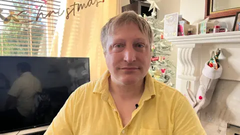 A man sits in front of a TV and white fireplace in a living room decorated for Christmas. He has short fair hair and is wearing a yellow shirt. A Christmas tree and window can be seen in the background. A gold "Christmas" sign hangs near the window. A stocking hangs from the fireplace.
