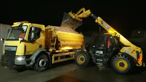 A yellow JCB vehicle uses its telescopic handler to load a yellow gritting vehicle with grit salt. Both vehicles are stationary in a dark depot. 